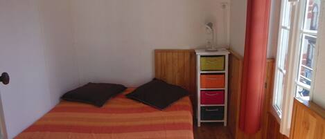 A small, single-bed room with a wooden floor, a bedside table with a lamp, and a window with curtains.