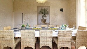 Un coin repas avec une longue table dressée pour six personnes, entourée de chaises en osier. Une plante en pot est suspendue au mur, au-dessus de la table.