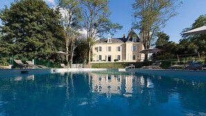 Une grande piscine entourée d’arbres, agrémentée de chaises longues et de parasols, et bordée d’un grand bâtiment au style architectural classique.