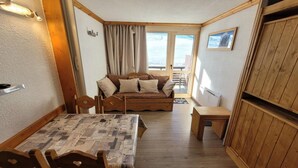 Ein gemütlicher Raum mit Esstisch und Stühlen aus Holz, einer Couch mit Kissen und einer kleinen Holzbank, mit Blick ins Freie durch ein Fenster.