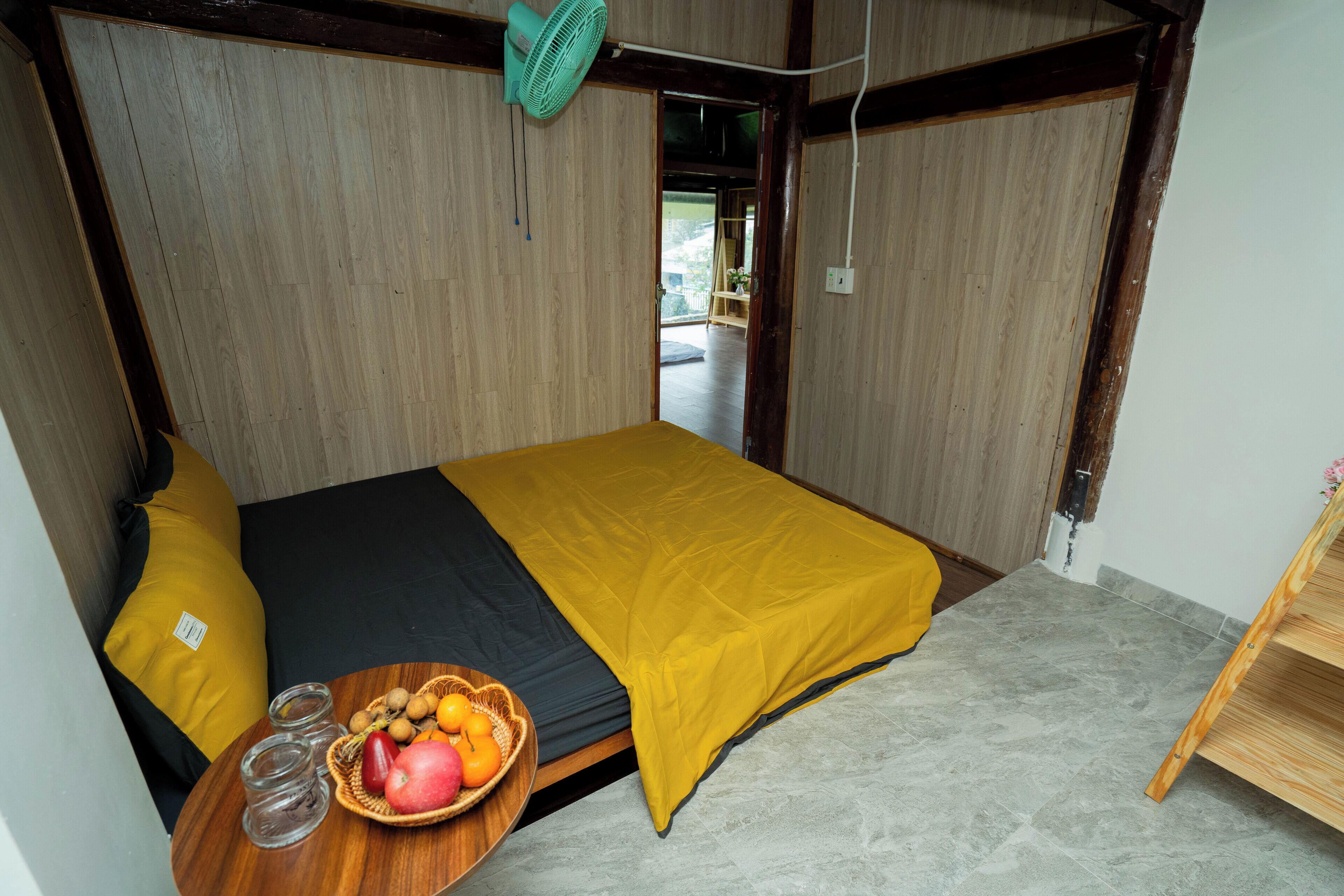 A room with a bed, a fan, a small table with a fruit bowl, and a view of the outside.