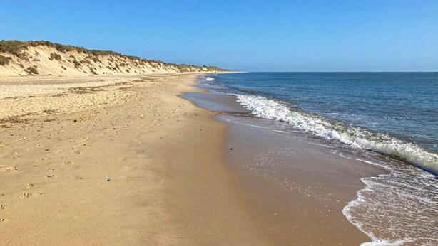 En bred, sandet strand med bløde bølger, klart himmel og en klit til venstre.