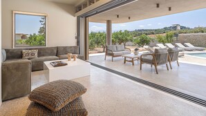 A modern living room with a sofa, coffee table, and wicker ottoman. There is a large window with a view of the outdoors. - Villa Portokalea, 200m From The Beach, Heated Pool (CHANIA)