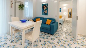 Un salón con sofá azul, mesa de comedor blanca y sillas. Hay un cuadro en la pared y una planta colgando del techo.