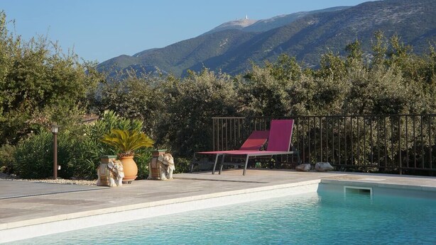 Une piscine avec un fauteuil de relaxation rouge et des plantes en pot, entourée d’une clôture et d’arbres, avec des montagnes en arrière-plan.