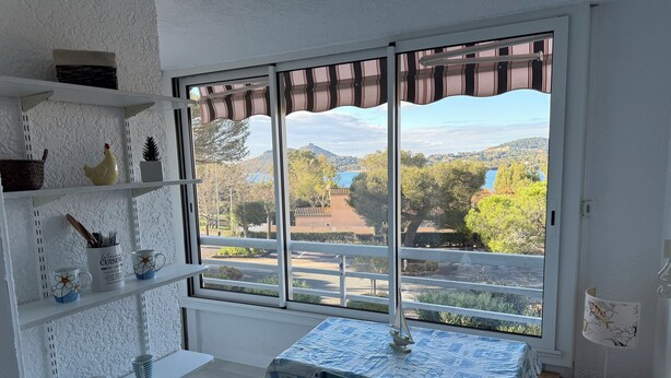 A room with a large window, a table with a patterned tablecloth, a chair, and shelves with various items. - Agay 4p, Piscine, Vue Mer, Tout a Pied (Saint-Raphaël)