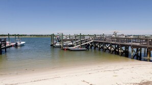 Eine Steg, der sich in ruhiges Wasser erstreckt, mit dort angelegten Booten, einem Sandstrand und einem wolkenlosen Himmel.