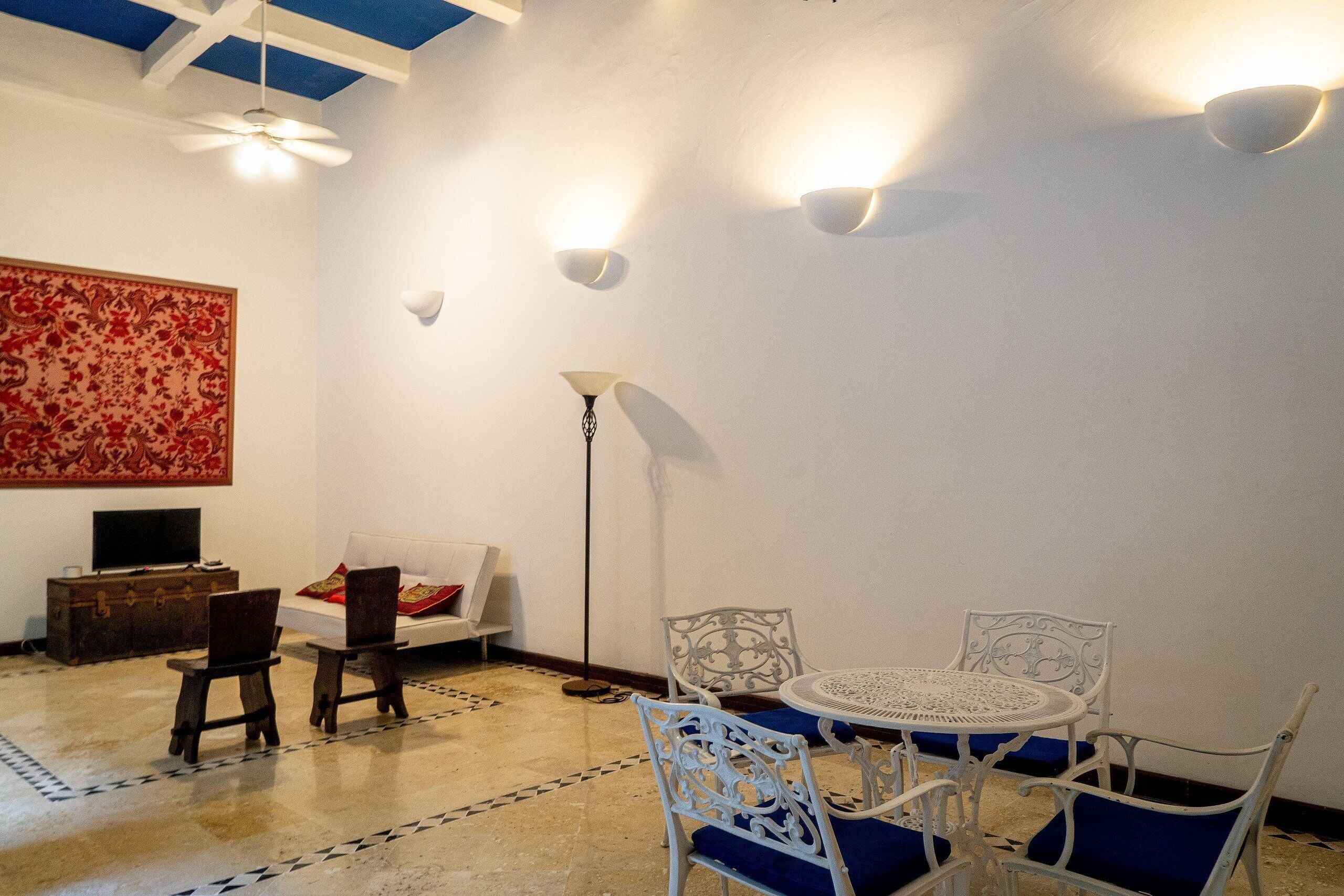 Une pièce avec un plafond blanc, une grande tapisserie rouge, un canapé blanc, une petite table en bois et une table ronde blanche avec des coussins bleus.