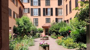 A courtyard with a central flower bed, surrounded by buildings with multiple windows and shutters. - Au Kugelhopf Doré - Historical Center (Colmar)
