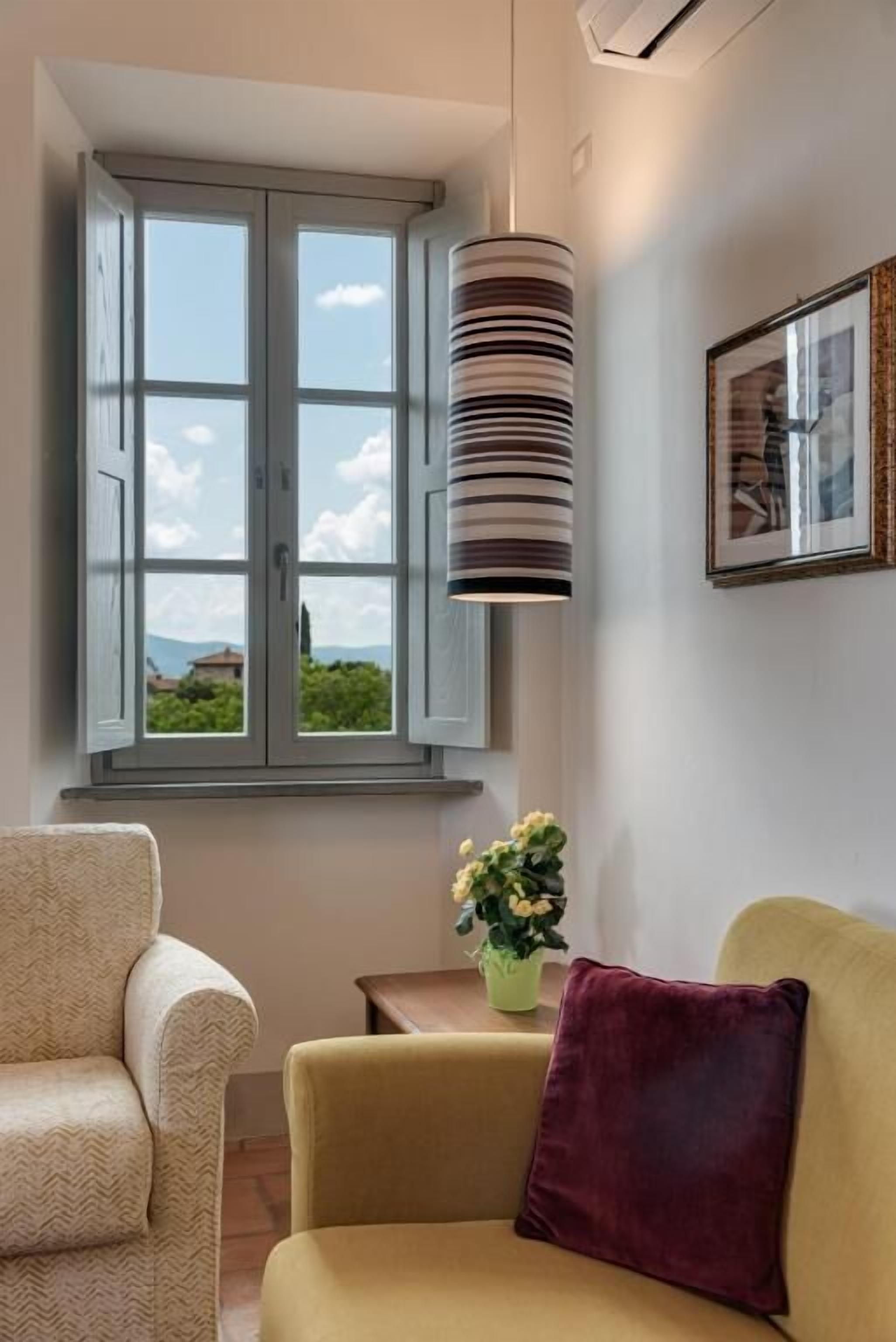 A room with a large window, a hanging lamp, a framed picture on the wall, and a small table with a plant.
