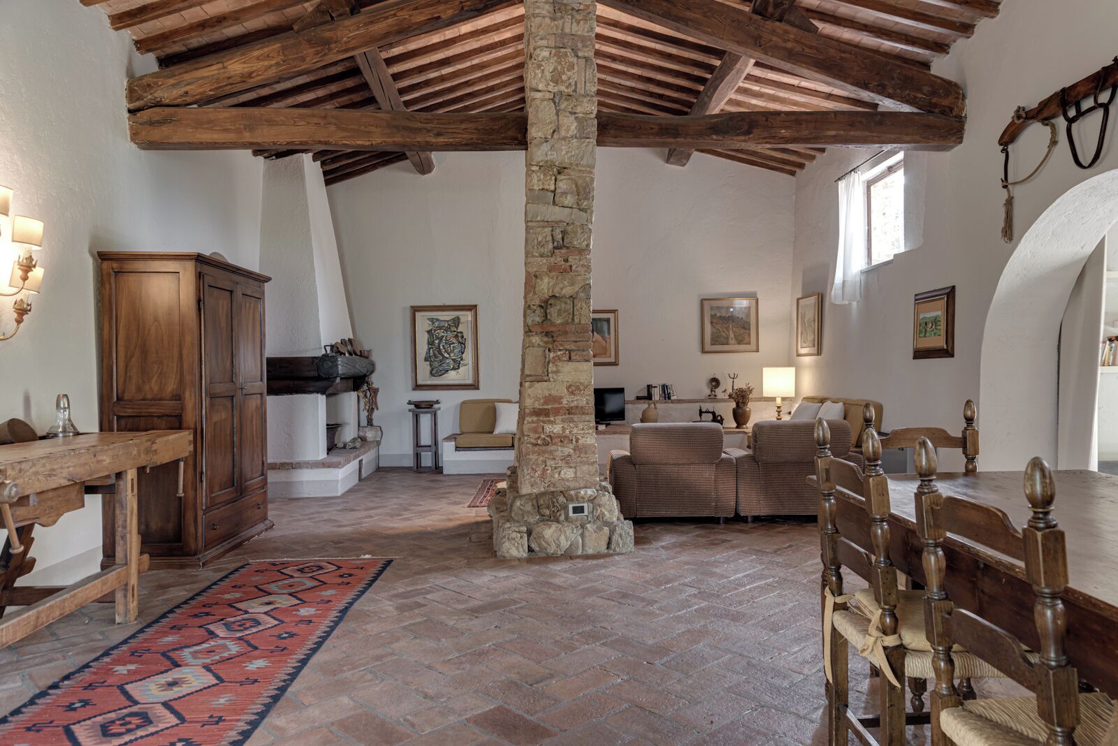 Ambiente rustico con travi a vista, una colonna in pietra, un tavolo in legno, un divano e un tappeto decorato.