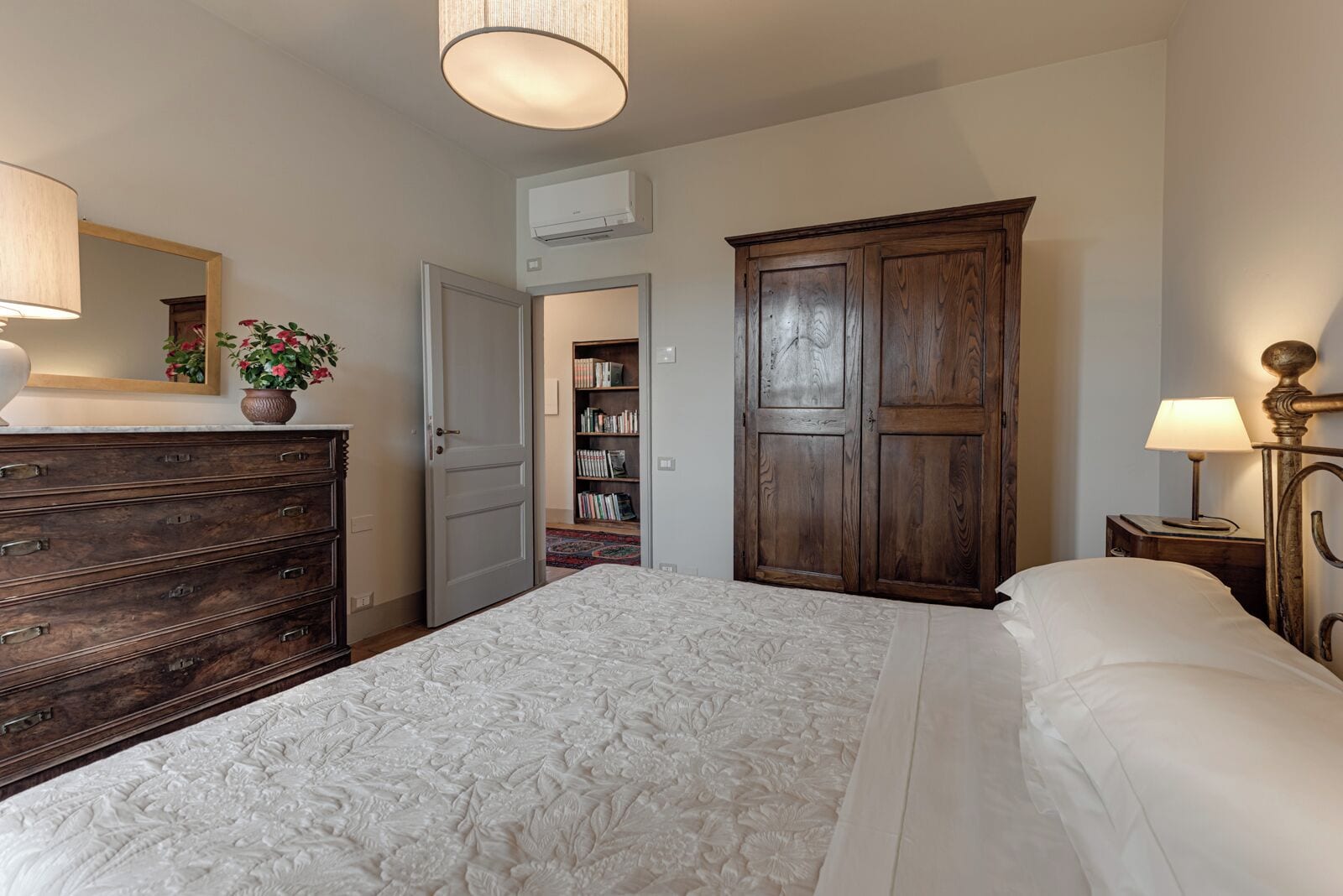 A bedroom with a large bed, a wooden wardrobe, a dresser, a mirror, and a bookshelf.