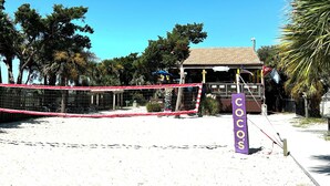 Ein Beachvolleyballfeld mit Netz, einem Schild mit der Aufschrift "COCOS" und einem Gebäude im Hintergrund.