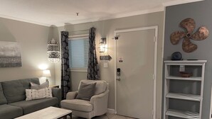 Ein Wohnzimmer mit grauem Sofa, einem kleinen Sessel, einem Holztisch, einer Wandregal und einer Tür mit Sicherheitsschloss.