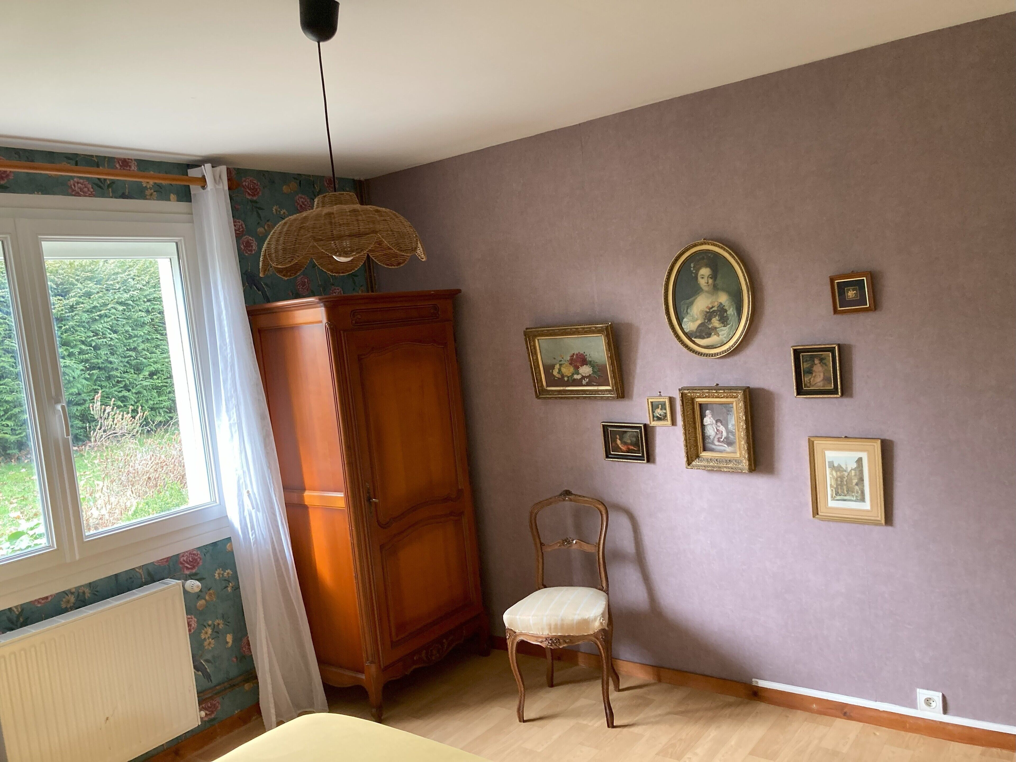 Una habitación con un ventanal grande, un armario de madera, una silla y varias fotografías enmarcadas en la pared.