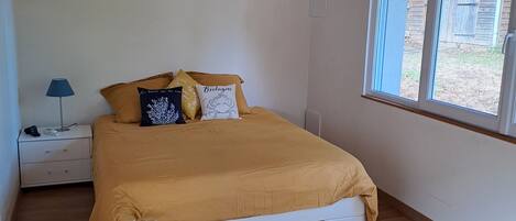 A bedroom with a wooden floor, a bed with a yellow bedspread, a nightstand with a lamp, and a window with a view of the outside.