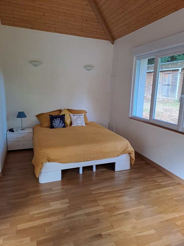 Un dormitorio con piso de madera, una cama con colcha amarilla, una mesita de noche con lámpara y una ventana con vista al exterior.
