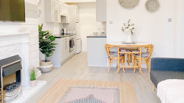 A modern kitchen with white cabinets, a dining area with a round table and wicker chairs, and a fireplace with a plant beside it.