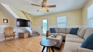 A living room with a wooden floor, a beige sofa, a round coffee table, a wooden chair, a staircase with a white railing, a ceiling fan, and a flat-screen TV mounted on the wall. - Charming 3-bedroom townhome 2 blocks from ocean and bay, in town of Stone Harbor (Stone Harbor)