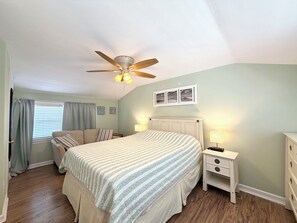 Une chambre à coucher comprenant un lit, une table de chevet, un ventilateur de plafond, une fenêtre avec des rideaux et des œuvres d’art murales.