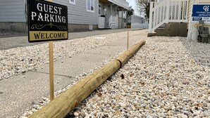 A guest parking area with a welcome sign, a wooden post, and a gravel path leading to a building with a balcony. - Charming 3-bedroom townhome 2 blocks from ocean and bay, in town of Stone Harbor (Stone Harbor)