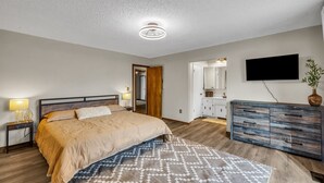A bedroom with a bed, bedside lamps, a dresser, a TV, and a door leading to a bathroom. - 4BR Retreat w/ KING Bed, Hot Tub & Prime Location (Owasso)