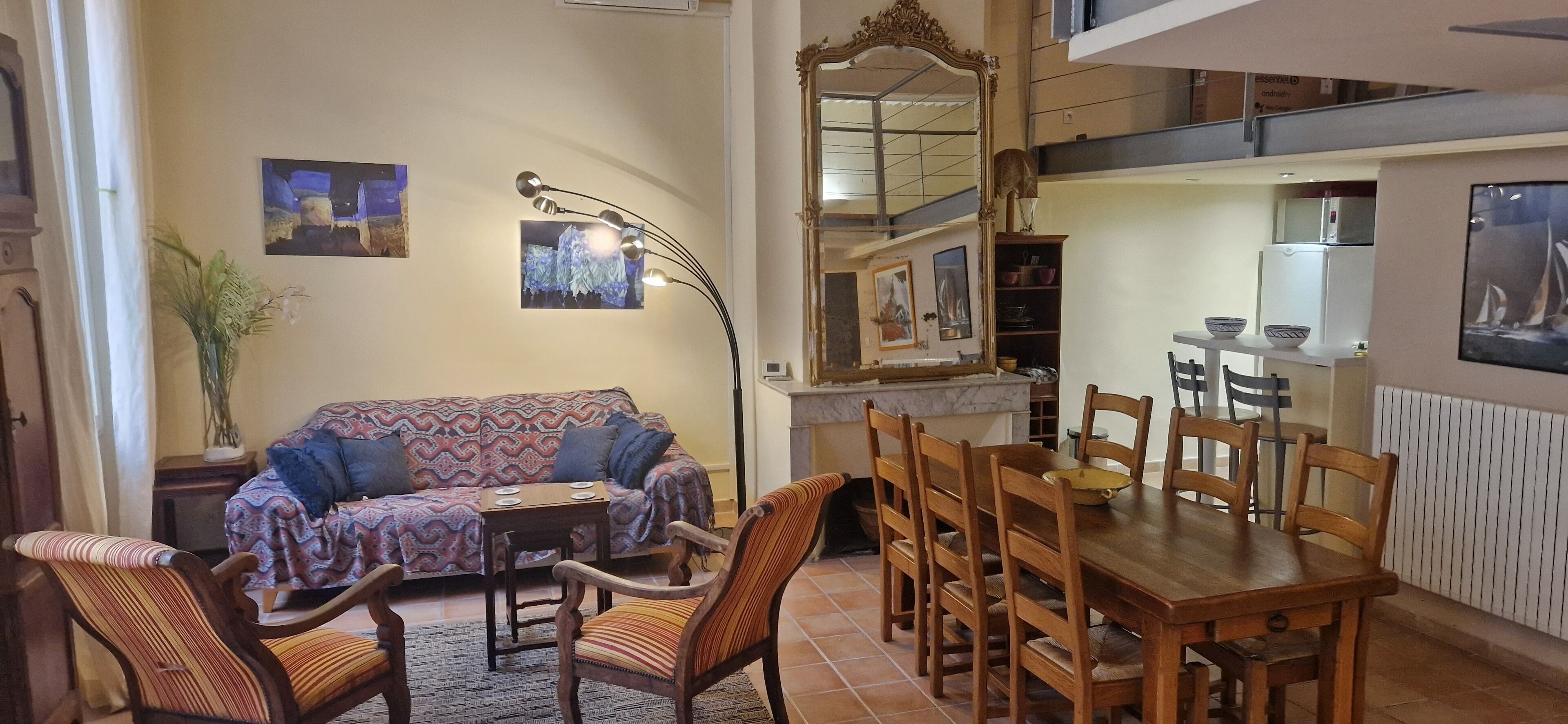 Ein gemütliches Wohnzimmer mit einem gemusterten Sofa, einem Holzesstisch und Stühlen. Über dem Tisch hängt ein großer Spiegel, und eine dekorative Lampe steht daneben.