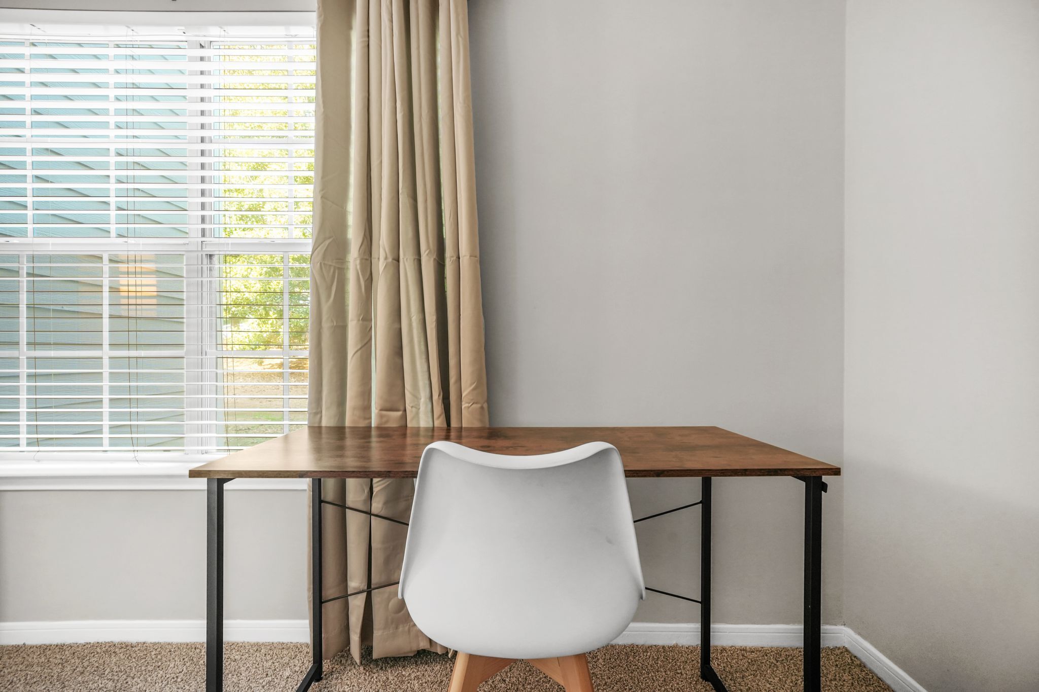 Een kamer met een houten tafel en een witte stoel, een raam met zonwering en een beige gordijn.