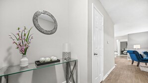 Un couloir avec une console en verre, un vase blanc contenant des fleurs roses, un miroir rond et une porte donnant sur une autre pièce.