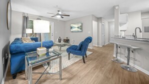 Un salon moderne avec un canapé bleu, une table basse en verre, un fauteuil bleu, un ventilateur de plafond et un coin cuisine doté d’un tabouret de bar.