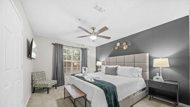 Une chambre à coucher comprenant un lit, des lampes de chevet, une chaise, un banc, un ventilateur de plafond et une fenêtre avec des rideaux.