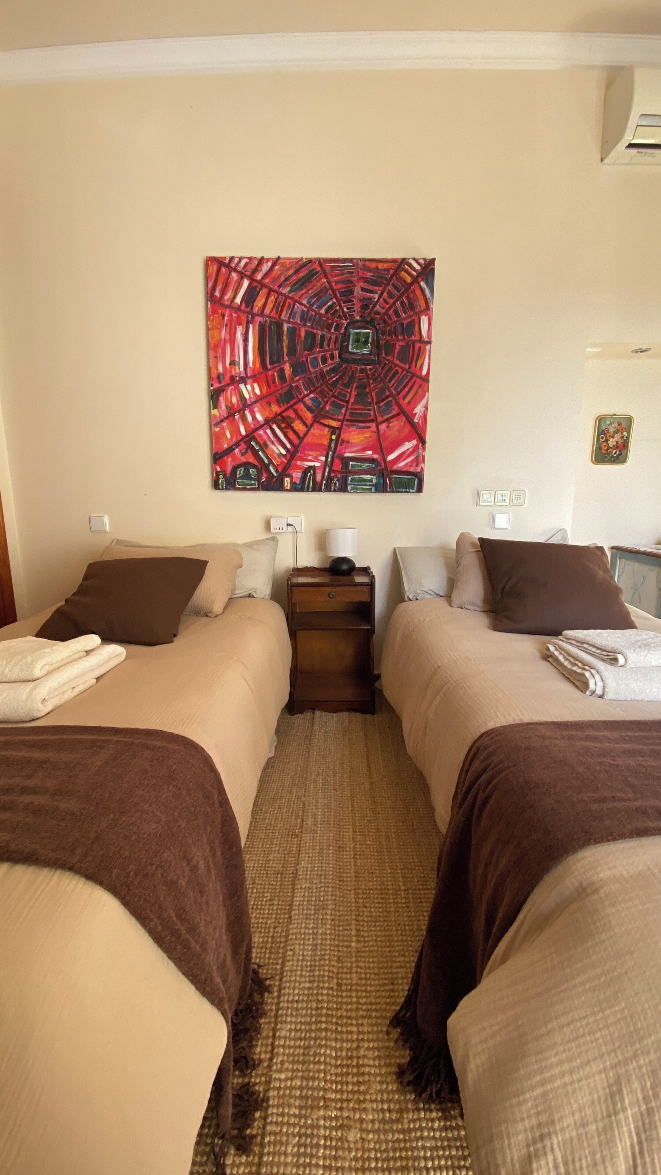 A hotel room with two beds, a large abstract painting on the wall, a small table with a lamp, and a woven rug.