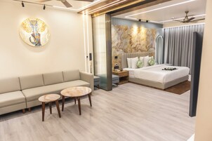 Una habitación de hotel moderna con sofá, dos mesas redondas, una cama con cabecera, una obra de arte grande en la pared y un ventilador de techo.