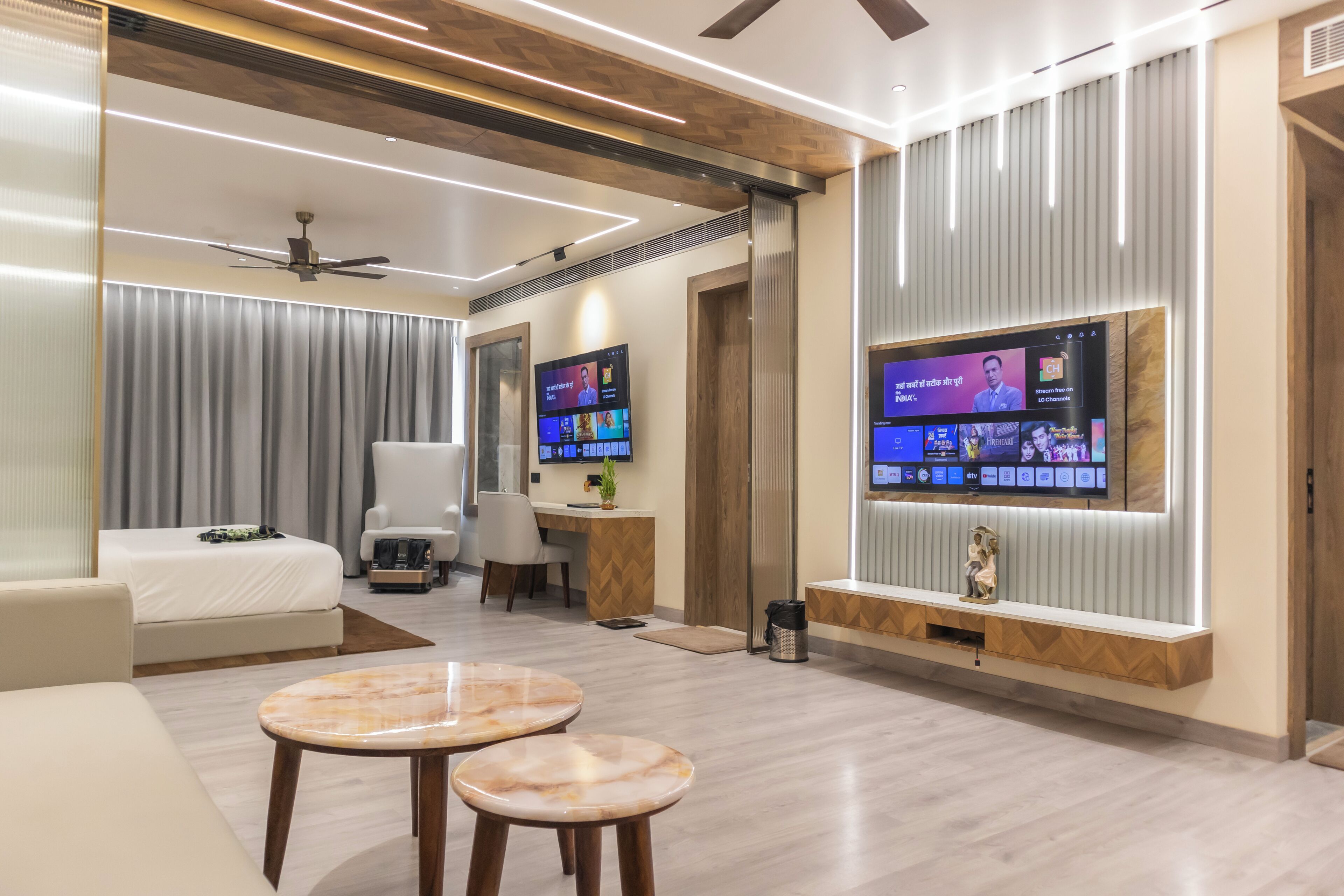 Une chambre d’hôtel moderne équipée d’un grand téléviseur à écran plat fixé au mur, d’un canapé, d’un bureau avec une chaise et d’une petite table avec deux tabourets.