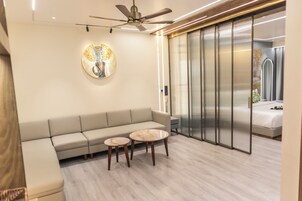 Una sala de estar moderna con un sofá, dos mesas de centro de mármol, un ventilador de techo y un ventanal corredizo que da a un dormitorio.