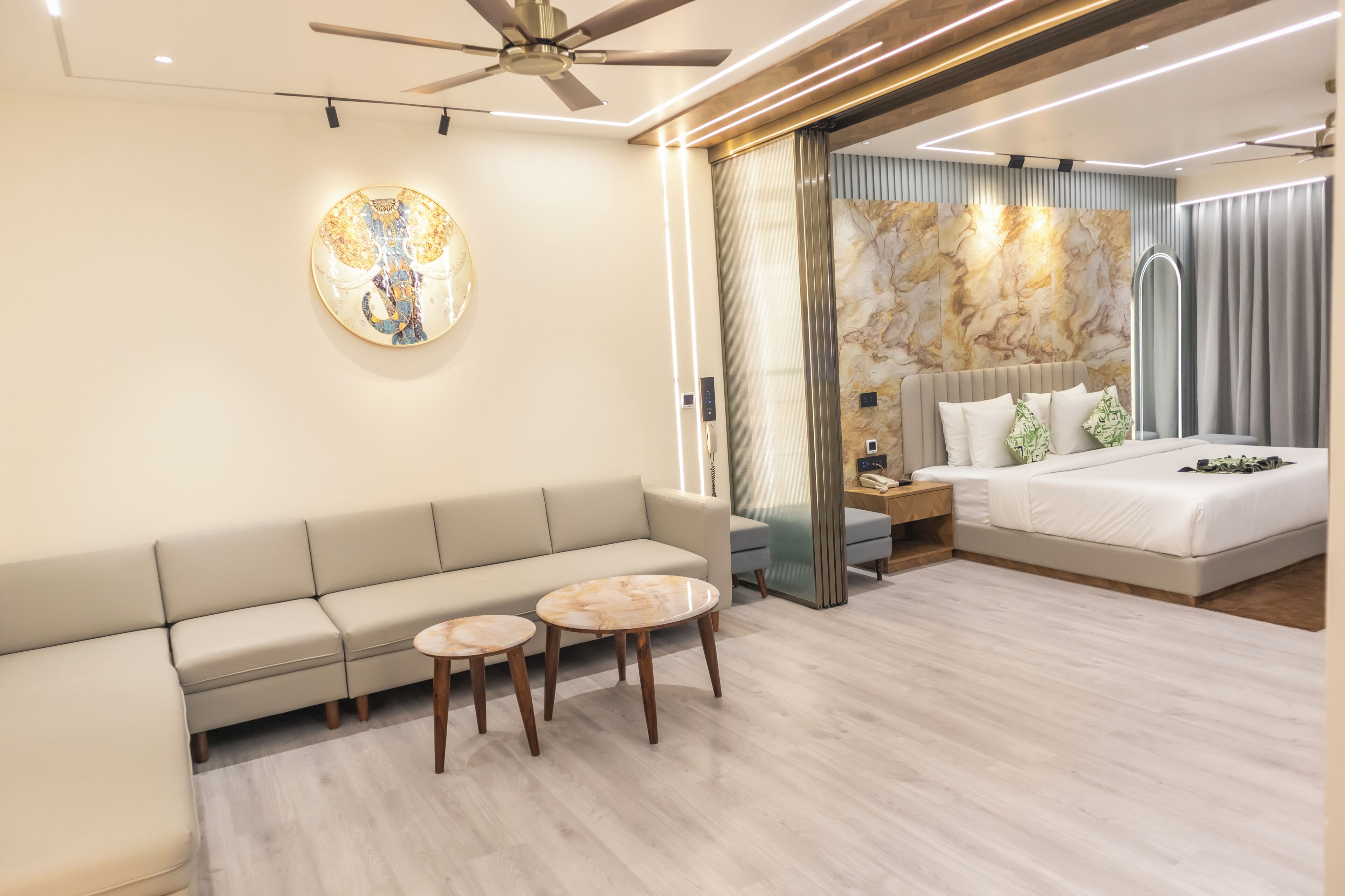 A modern hotel room with a sofa, two round tables, a bed with a headboard, and a ceiling fan.