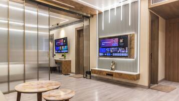 Een moderne hotellobby met een grote flatscreen televisie bevestigd aan een houten paneel, een receptiebalie en twee tafels met marmeren blad.