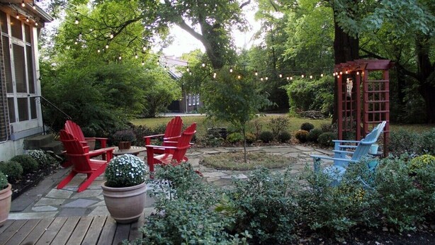 Una terraza de madera con sillas Adirondack rojas y azules, una pérgola con luces colgantes y un jardín con diversas plantas.
