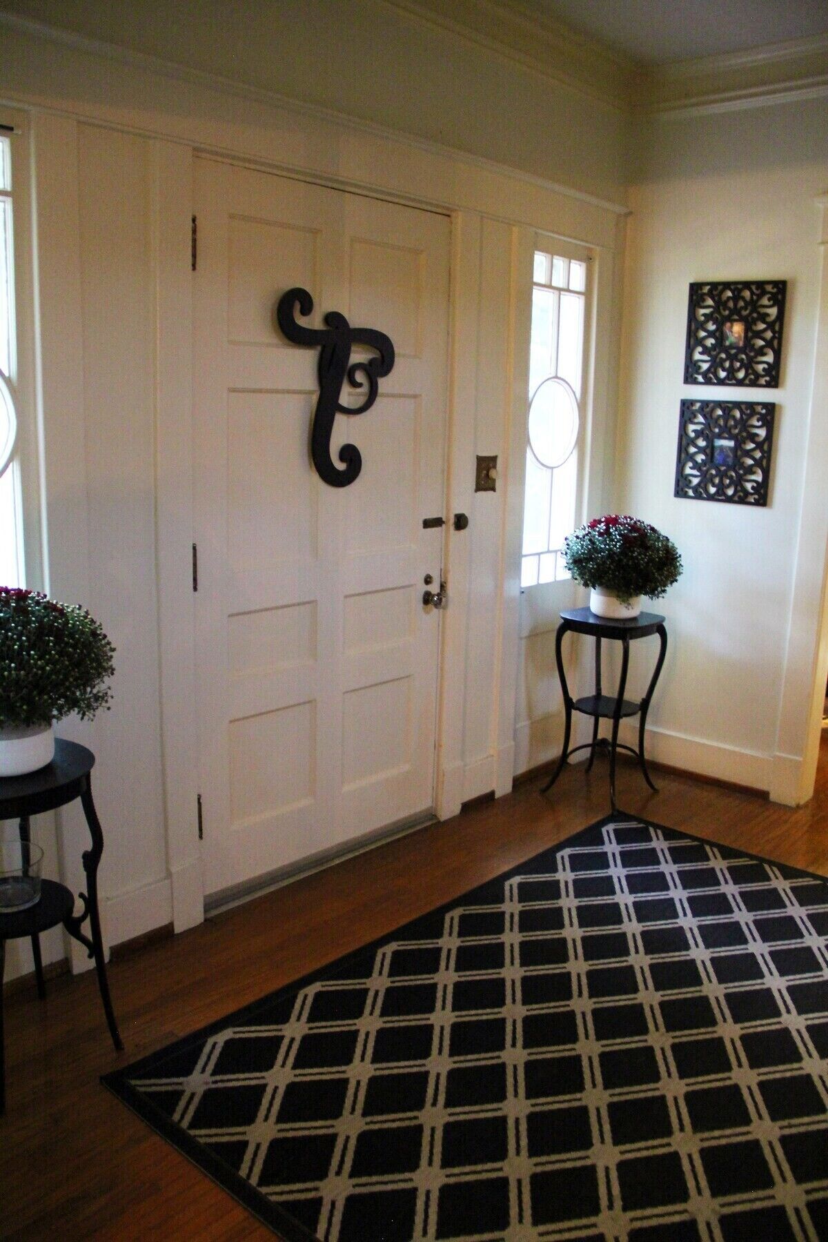 A hallway with a black and white checkered rug, a white door with a black decorative letter, and two potted plants on side tables.