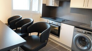 A compact kitchen with a stove, oven, and washing machine, a window with blinds, and three black chairs at a table. - 3br-sleeps5-balcony-centrallondon-ws95 (London)