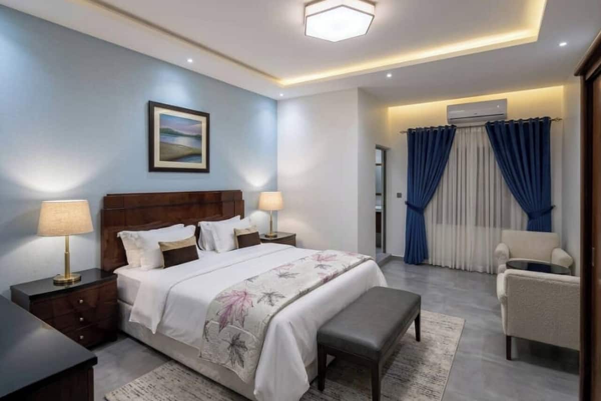 A bedroom with a bed, bedside tables, a bench, a chair, and a view of a balcony with curtains.