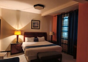 A hotel room with a bed, bedside lamps, a nightstand, a chair, a window with curtains, and a painting on the wall. - AMSY HOTEL AMSY HOTEL (Dakar)