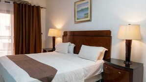 A hotel room with a bed, two bedside lamps, a nightstand, a painting on the wall, and an air conditioning unit. - AMSY HOTEL AMSY HOTEL (Dakar)