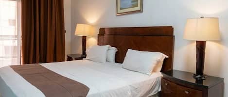 A hotel room with a bed, two bedside lamps, a nightstand, a painting on the wall, and an air conditioning unit.