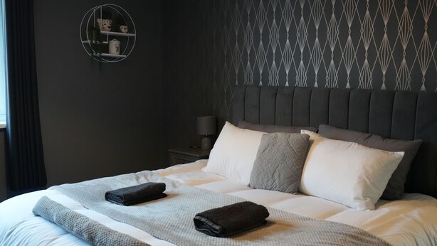 Cama bem arrumada com um edredom cinza e branco, duas toalhas enroladas e um cobertor cinza.