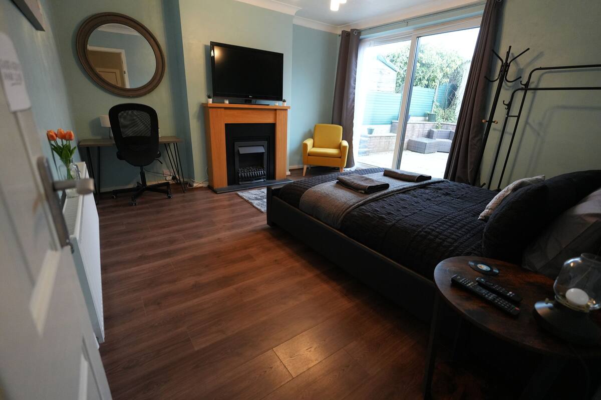 A bedroom with a bed, a TV mounted above a fireplace, a chair, and a desk.