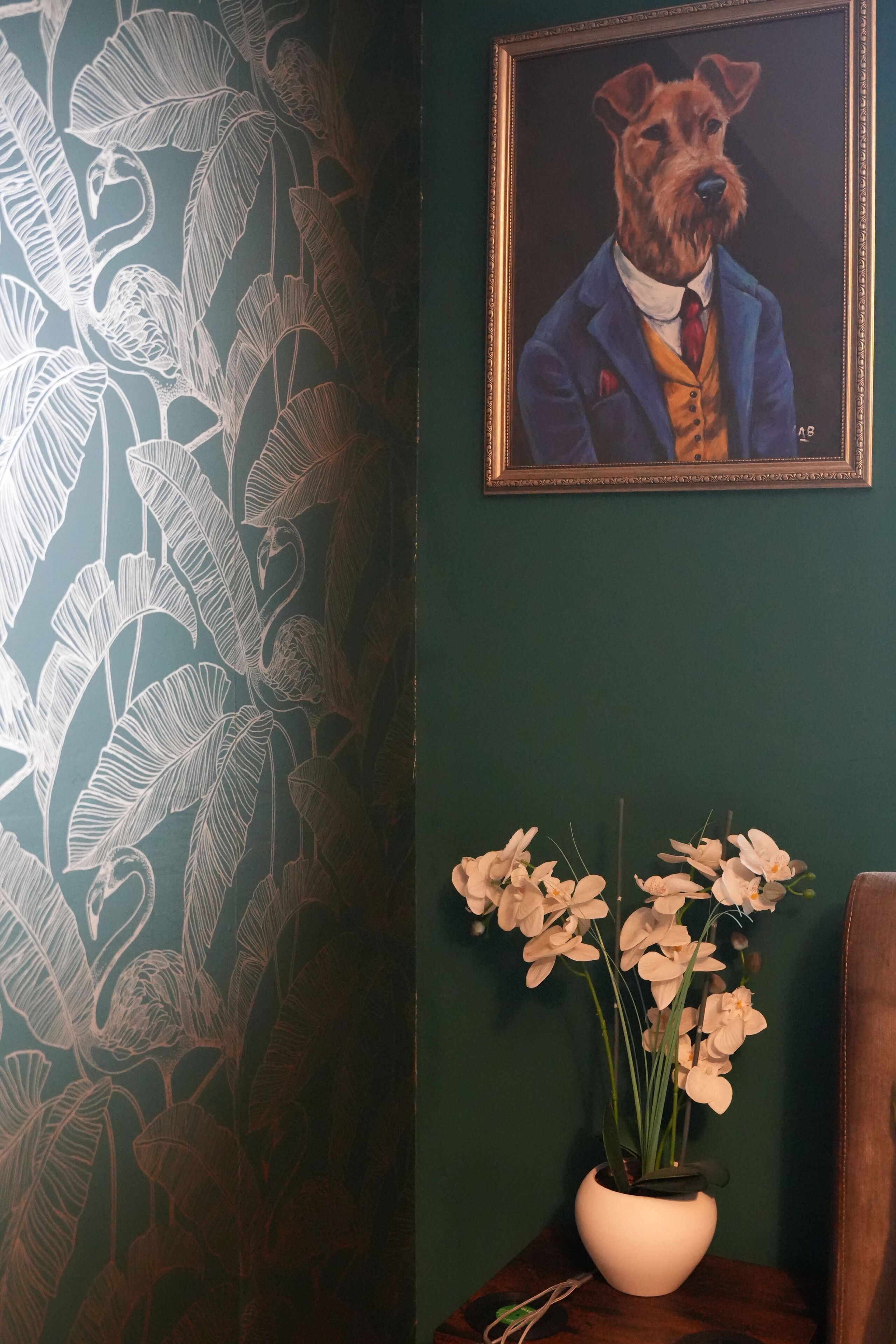 A wall with a framed painting of a dog in a suit and a vase of orchids.