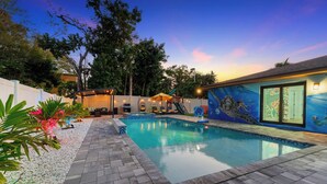 A swimming pool with a tiled deck, surrounded by a white fence and lush greenery.