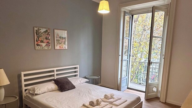 A bedroom with a bed, a bedside table, a lamp, a ceiling light, and a window with a view of trees.
