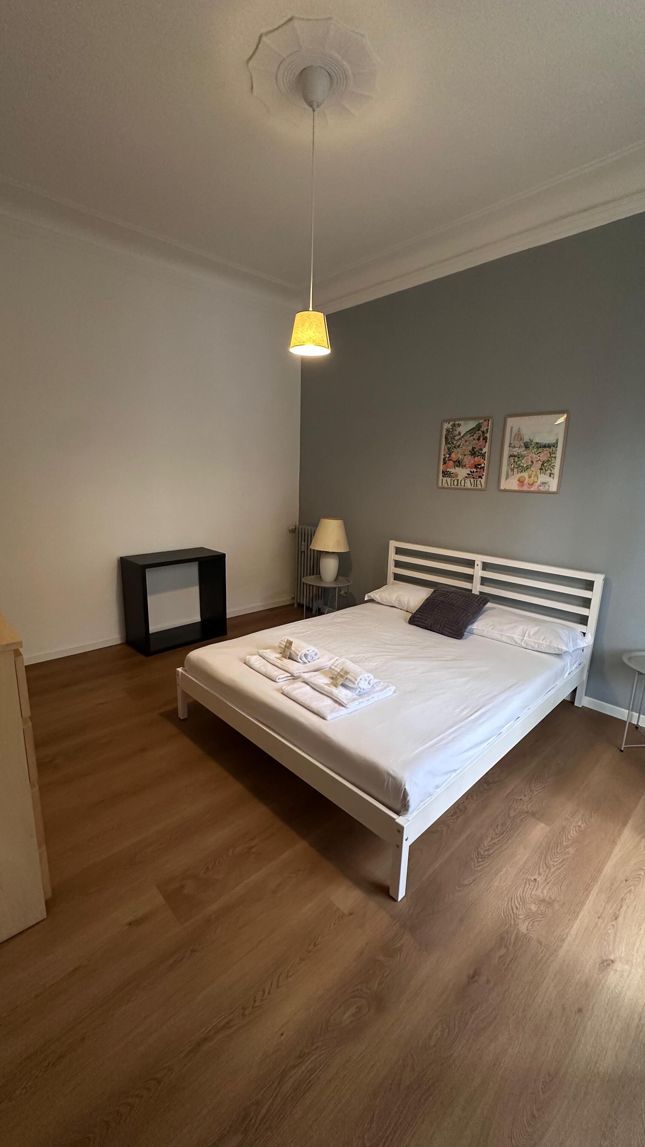 Une chambre à coucher bien rangée, avec un lit, une table de chevet, une lampe et une œuvre d’art accrochée au mur.